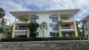 A modern, multi-story white apartment building or resort with spacious balconies, yellow support pillars, and palm trees in front under a cloudy sky.
