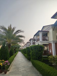 A landscaped pathway lined with neatly trimmed hedges and flowering plants leads through a residential area featuring two-story houses with balconies. 
