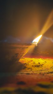 A close-up of a traditional oil lamp with a flickering flame, set against colorful yellow and pink powder.