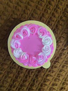 A round pink cake with white and pink swirled frosting rosettes on top, set on a textured brown surface, creating a festive and sweet atmosphere.