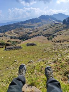 A person’s feet in hiking shoes are positioned on a rocky outcrop, overlooking a vast, rolling landscape of green hills and winding roads.
