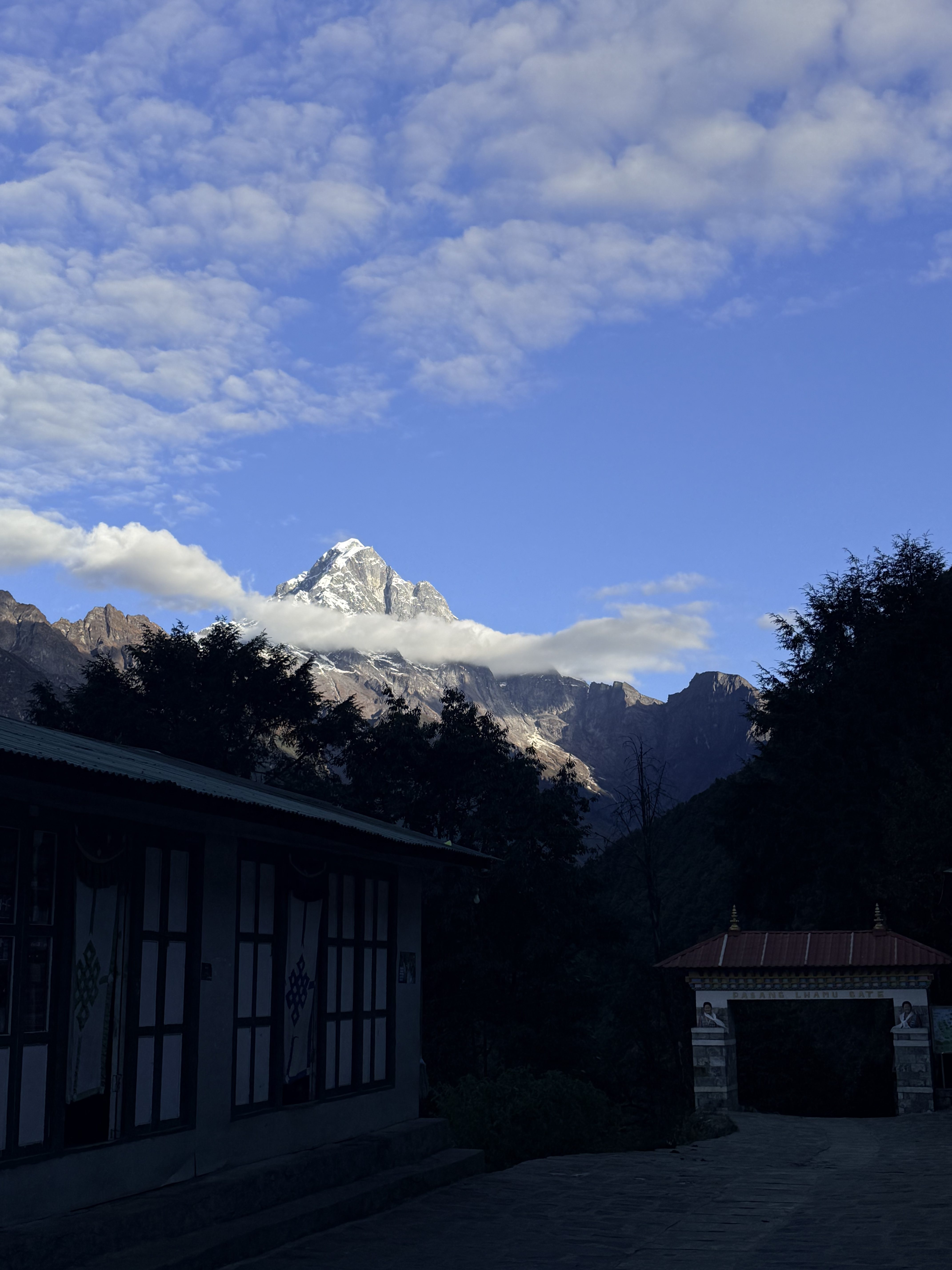 A scenic view of a snow-capped mountain peak under a blue sky with scattered clouds. In the foreground, there is a building with large windows and traditional decorative elements, along with a rooftop covered in color.