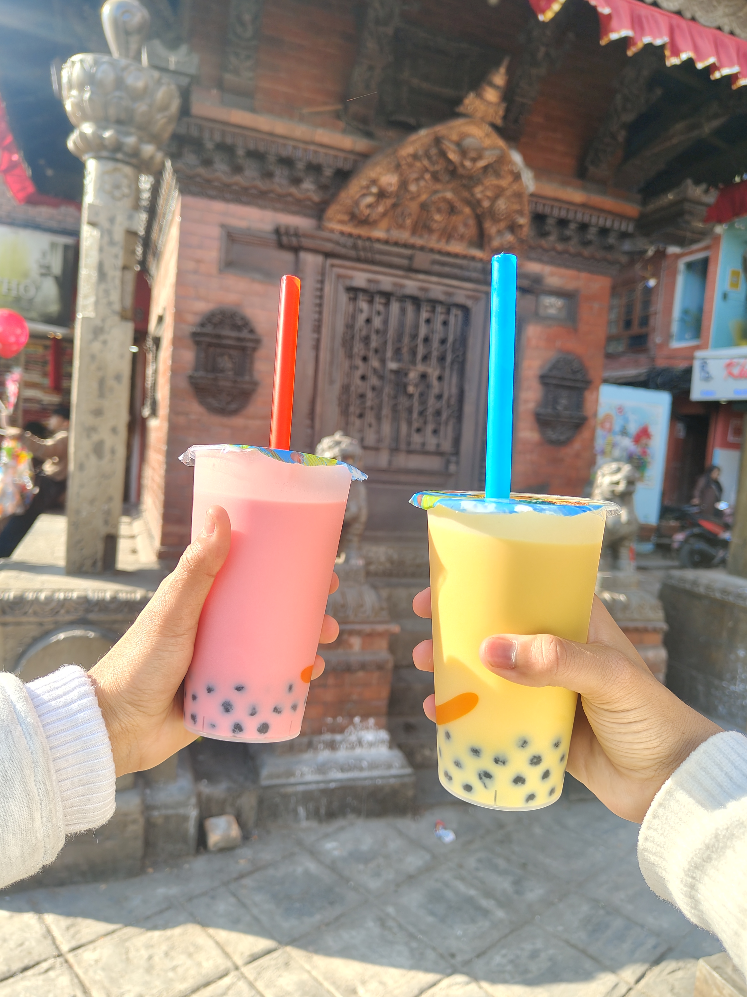 Two hands are holding colorful bubble tea drinks&mdash;one pink with red straw and the other yellow with blue straw. 