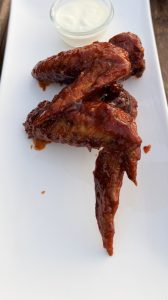 
A plate featuring three glazed chicken wings, coated in a shiny red sauce, arranged in a dynamic, overlapping position. In the background, there is a small bowl of creamy dipping sauce.