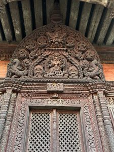 A detailed close-up of ornate wooden carvings on a temple entrance. The central panel features intricate designs including a figure surrounded by various mythical creatures and floral patterns