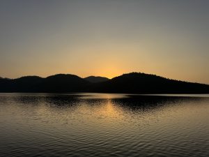 A serene lake at sunset, with smooth water reflecting the soft, warm glow of the sun as it dips behind dark, rolling hills in the background