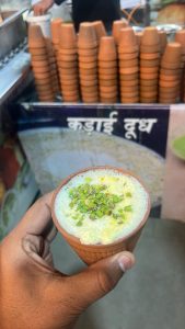 A person holds a small clay cup of frothy, light green Jodhpur-famous kadhai dudh topped with finely chopped pistachios.

