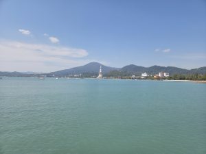 A calm blue bay with small boats, green hills in the background, and a tall white lighthouse near the shore surrounded by buildings and greenery.