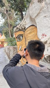 A person is standing on a pathway, facing a large rock wall that features a painted face. The face is stylized with prominent features, including a large eye and a slight smile, in warm yellow and orange tones.