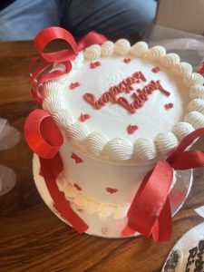A round white cake topped with a decorative border of whipped cream and red ribbon accents. The cake is adorned with small red heart-shaped decorations and features the phrase "happy birthday" written in red icing on the top.