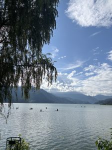 A serene lakeside scene featuring several kayakers paddling on calm waters. In the foreground, lush green foliage partially obscures the view, while in the background, majestic mountains rise under a blue sky dotted with fluffy white clouds. 
