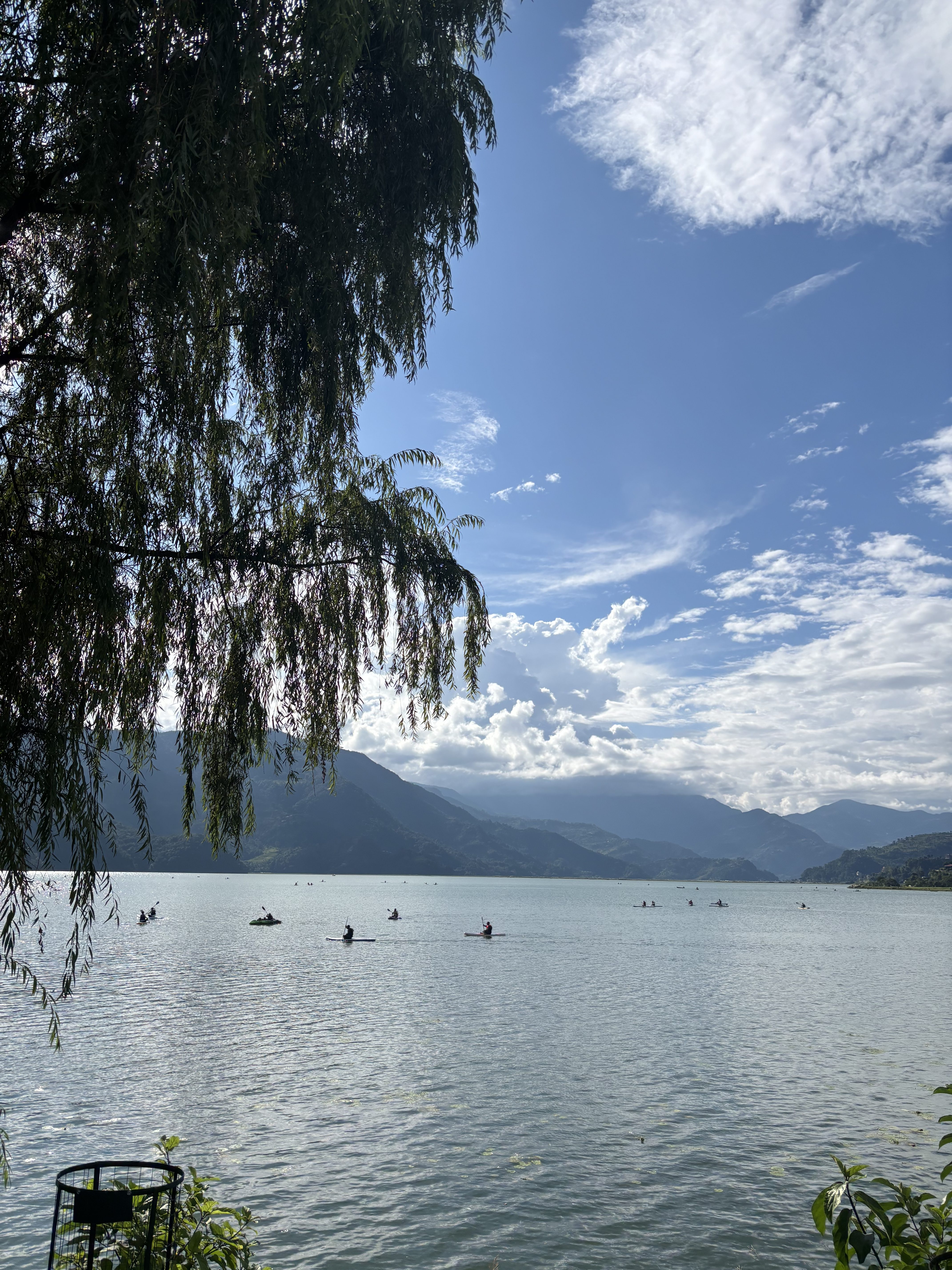 A serene lakeside scene featuring several kayakers paddling on calm waters. In the foreground, lush green foliage partially obscures the view, while in the background, majestic mountains rise under a blue sky dotted with fluffy white clouds. 