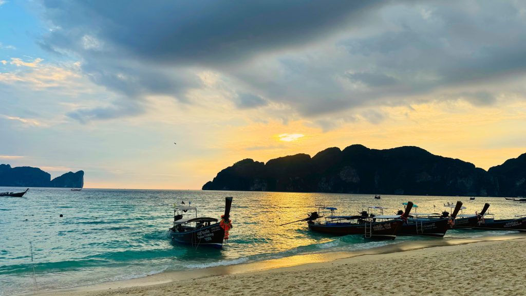 Long-tail boats moored near a sandy beach at sunset with calm sea and dark limestone islands on the horizon view at Phi Phi Island, Thailand.