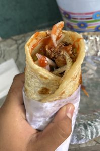 A hand holding a rolled wrap filled with cooked meat, sliced onions, and colorful sauces, wrapped in a thin, soft flatbread. 