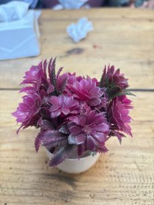 A cluster of vibrant pink artificial flowers with green leaves, arranged in a small, white pot. 
