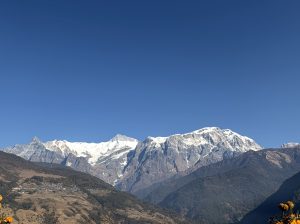 A panoramic view of snow-capped mountains under a clear blue sky, with a valley in the foreground featuring hills and patches of greenery. 