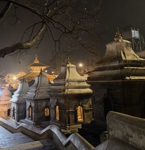 A peaceful nighttime scene featuring a series of intricately designed temples or shrines with distinct curved roofs and ornate decorative elements. In the background, more structures are faintly visible among soft lighting. Cobblestone pathways lead through the area, enhancing the serene atmosphere.