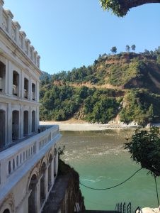 A serene landscape featuring a partially visible white stone building with arches on the left side, overlooking a river with calm, greenish water. 