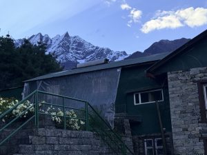 A green stone building with flower-lined stairs in front, set against snow-capped mountains and a blue, partly cloudy sky.
