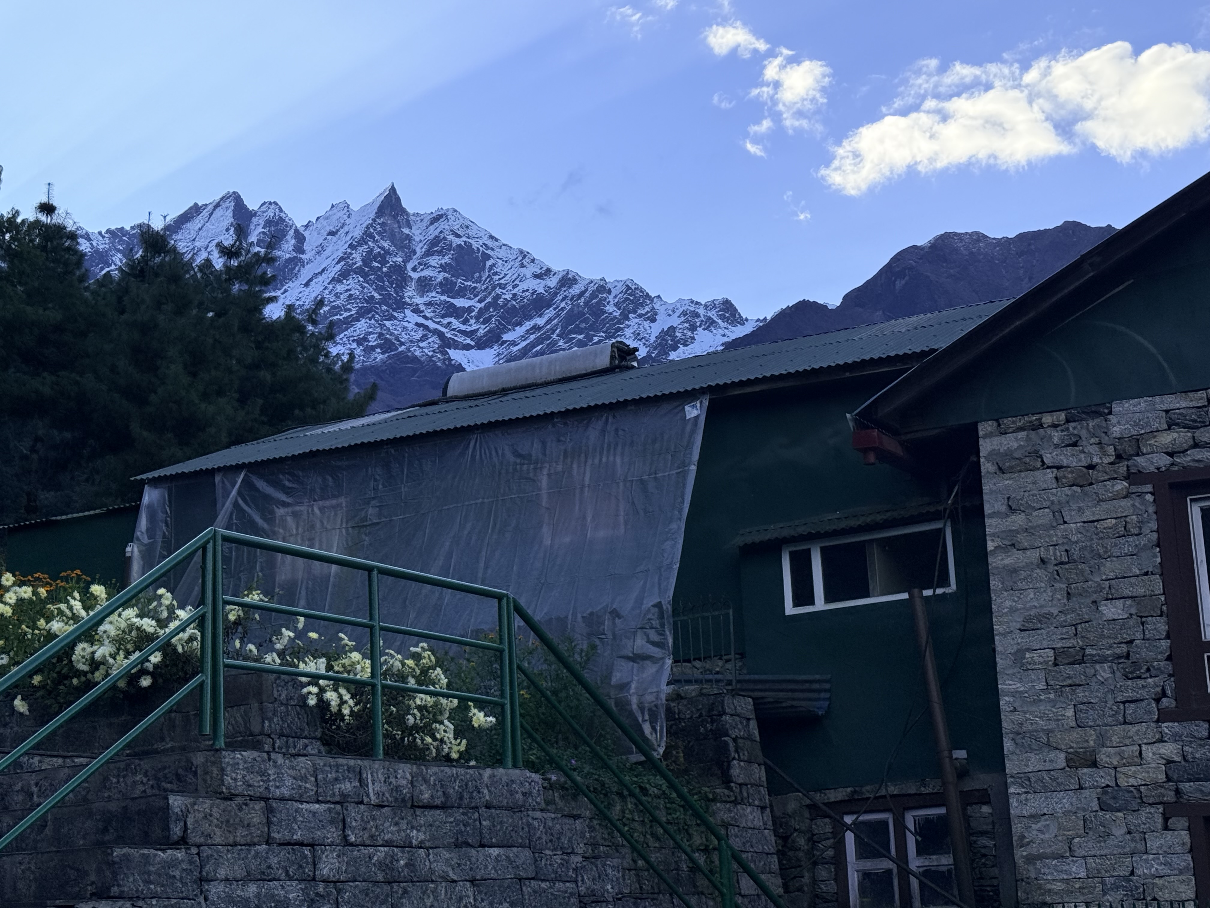 A green stone building with flower-lined stairs in front, set against snow-capped mountains and a blue, partly cloudy sky.
