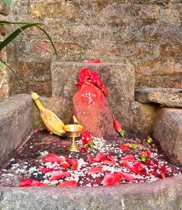 A stone altar adorned with vibrant red flower petals and small white rice grains. A small oil lamp is lit, illuminating the space.