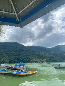 
A serene lakeside scene featuring several colorful wooden boats moored on the water. In the background, lush green hills rise beneath a cloudy sky, creating a tranquil atmosphere.