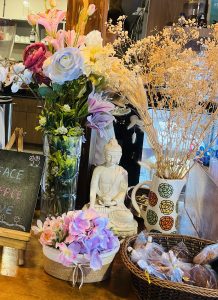 A cozy display featuring a mix of faux flowers in varied colors including pink, purple, and white, arranged in a tall glass vase. In the center, there is a white Buddha statue sitting peacefully