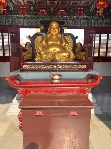 A large golden Laughing Buddha statue sits on an ornately carved wooden platform, smiling cheerfully.
