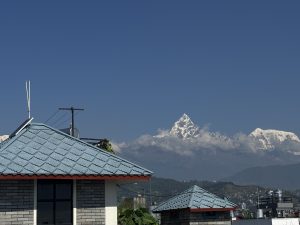 Snow-capped Machapuchre rises under a clear blue sky, with two buildings in the foreground—one with a blue roof and the other brick with a window.