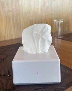 A small square tissue box with a crumpled paper tissue partially sticking out of the top, placed on a textured brown table surface.