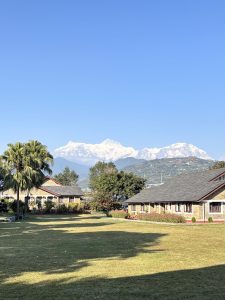 A scenic outdoor view featuring a green lawn and several buildings with stone facades and sloped roofs, surrounded by trees and plants. In the background, majestic snow-capped mountains rise against a clear blue sky, creating a picturesque landscape.