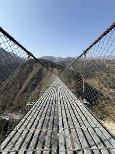 
A suspension bridge stretches across a valley, with a wooden plank surface and metal railing. 