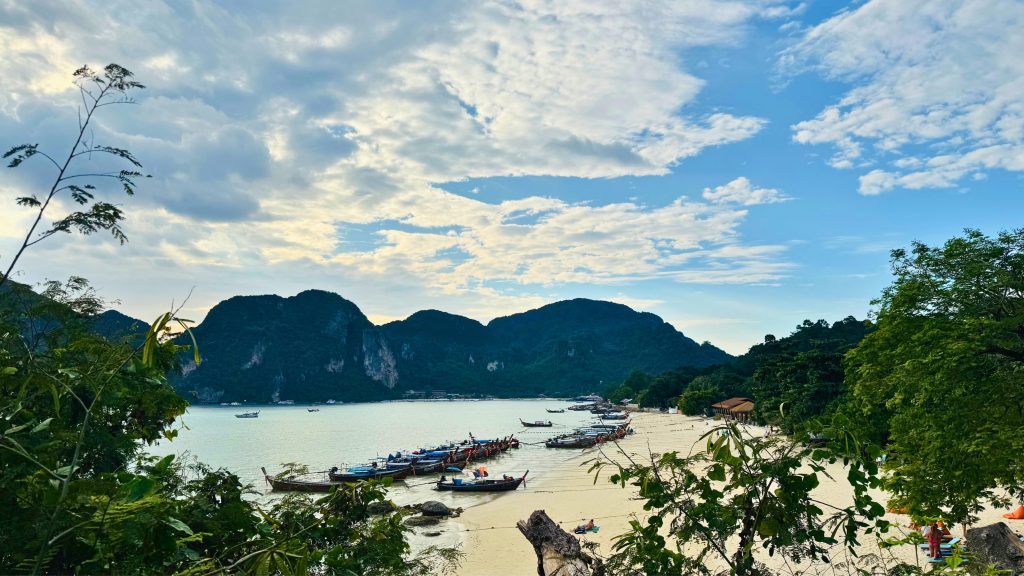 Long-tail boats anchored along a sandy beach with turquoise water and limestone cliffs under a partly cloudy sky at Phi Phi Island, Thailand.