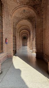 A long corridor with a series of red brick arches overhead, featuring wooden doors along one side and smooth concrete flooring. 
