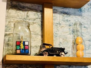 A wooden shelf holding a glass jar with a Rubik's cube inside, a small metal toy car, and a tall bottle filled with orange balls.