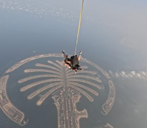 Aerial view of two skydivers descending from a height over a large, palm-shaped island surrounded by blue water.