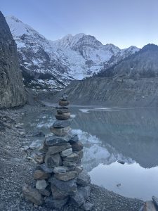A tall stack of flat stones stands on a rocky lakeshore. Behind it, a calm, icy-blue lake reflects steep, snow-covered mountains and dark cliffs in Gangapurna Lake, Manang, Nepal.
