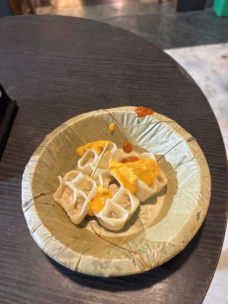 A small, round, green disposable plate contains several dumplings filled with meat, topped with a yellow sauce and small dollops of a reddish sauce.