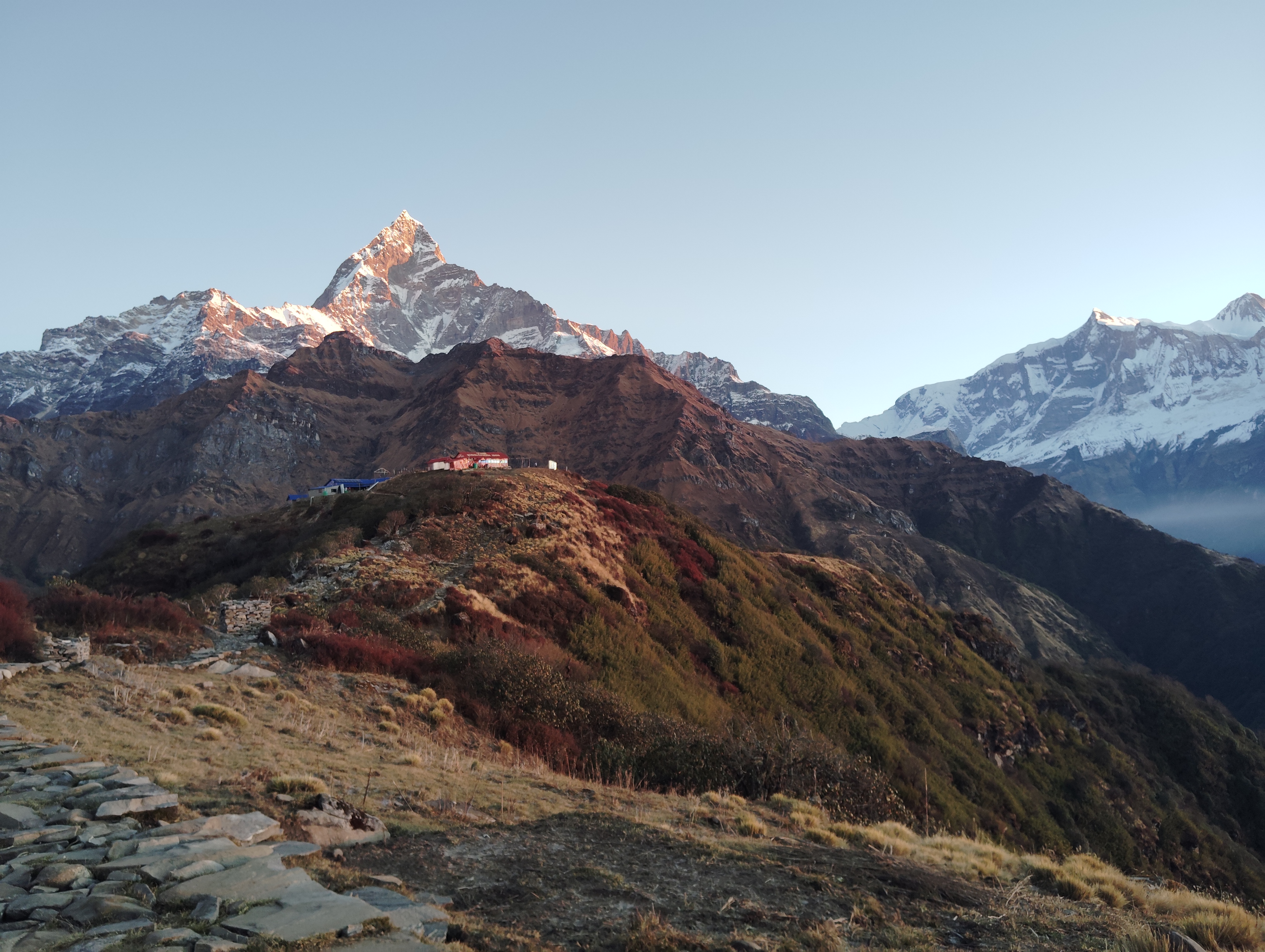 A panoramic view of majestic mountains, featuring a snow-capped peak illuminated by the sun, with rugged terrain and rocky pathways leading to a small settlement nestled on a hill.