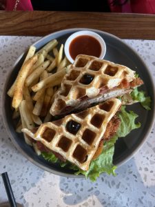 
A plate featuring a waffle sandwich cut in half, filled with lettuce, a slice of tomato, and possibly turkey or chicken, garnished with a single olive on to