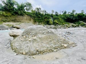 Large rocks lie near a flowing Seti Gandaki river with green cliffs and a small waterfall in Pokhara, Nepal, showing raw nature and earthy textures.