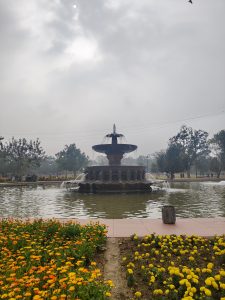 A decorative fountain stands in the center of a pond surrounded by flowers. In the foreground, vibrant orange and yellow marigolds bloom alongside the water's edge, while the fountain features tiered levels with water flowing over the sides.