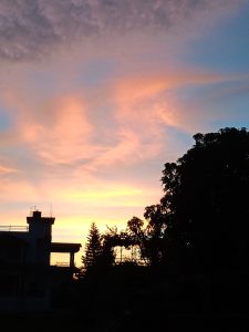 A silhouette of a house with a visible rooftop and antenna is in the foreground against a colorful sunset sky.