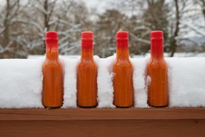 Four bottles of orange habanero hot sauce with red wax sealed lids sitting on a snow-covered railing with snow-covered tree branches in the background.
