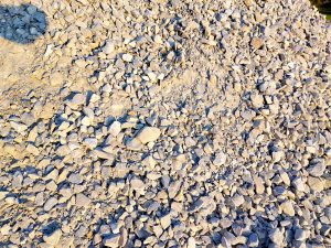 A close-up view of a pile of small, irregularly shaped gray rocks and pebbles scattered across a sandy surface, with varying textures and sizes, illuminated by sunlight.