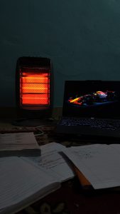 A warm, glowing electric heater is positioned on the left side of the image, casting a reddish light. To the right, a laptop displays a visually striking image of a racing car against a dark background. On the floor, there are scattered study materials, including notebooks with handwritten notes and printed papers, all resting on a patterned rug.