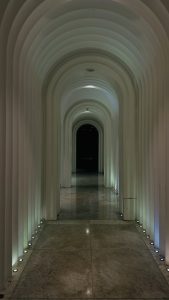 The image depicts a modern, well-lit corridor characterized by a series of arches along its sides. The walls are designed with rounded, curved elements, and the floor features polished stone tiles.