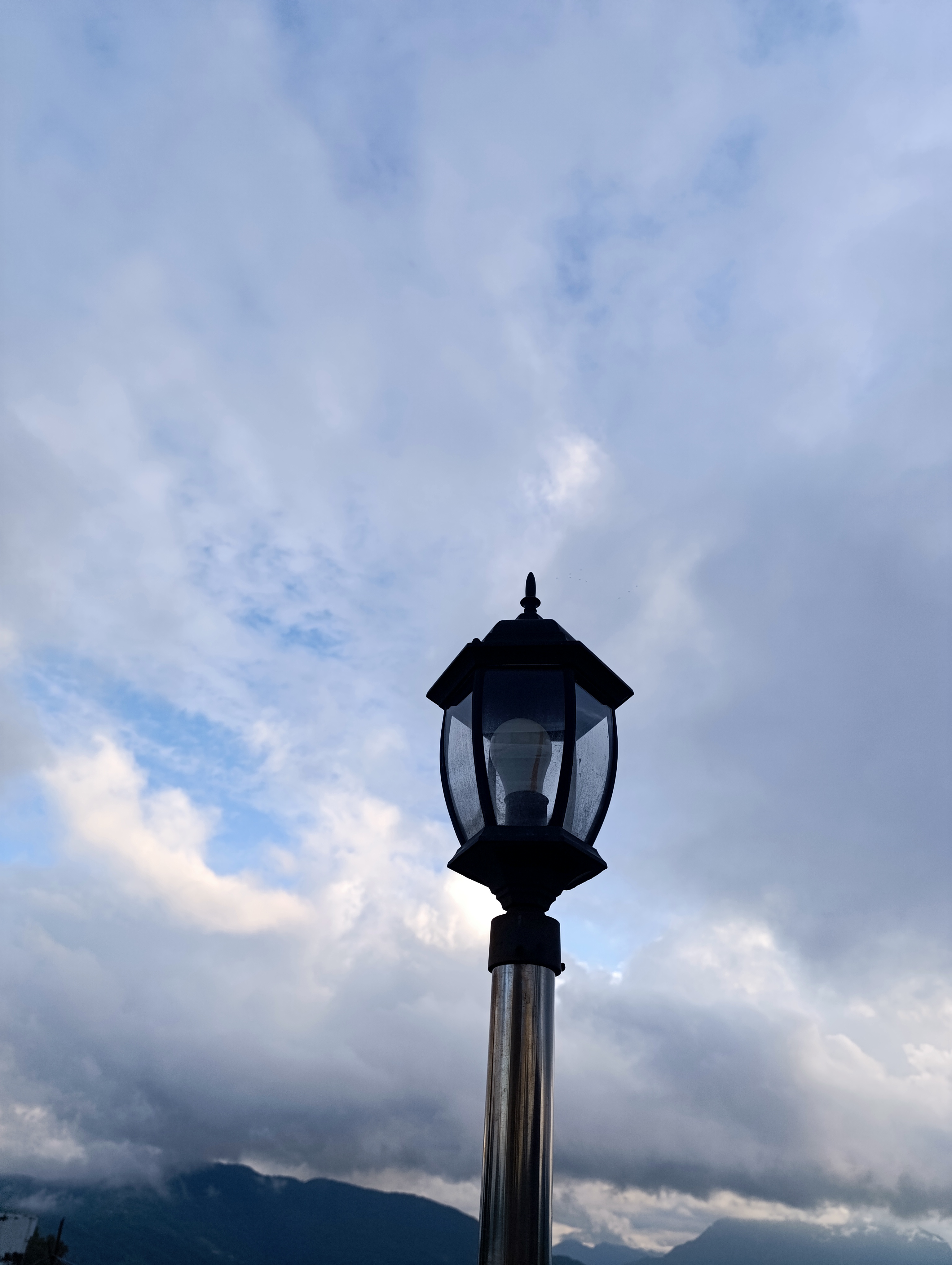 The top of a street lamp on a post, set against a a partly cloudy sky.