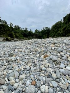 A rocky riverbed filled with a variety of smooth stones in shades of gray and beige, surrounded by lush green trees and cliffs under a cloudy sky in Pokhara, Nepal. The scene captures a serene, natural landscape with a hint of moisture in the air.
