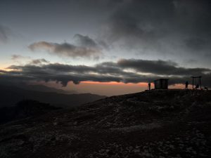 A silhouette of two figures standing on a hilltop at dusk, with a small wooden structure and a swing in the background